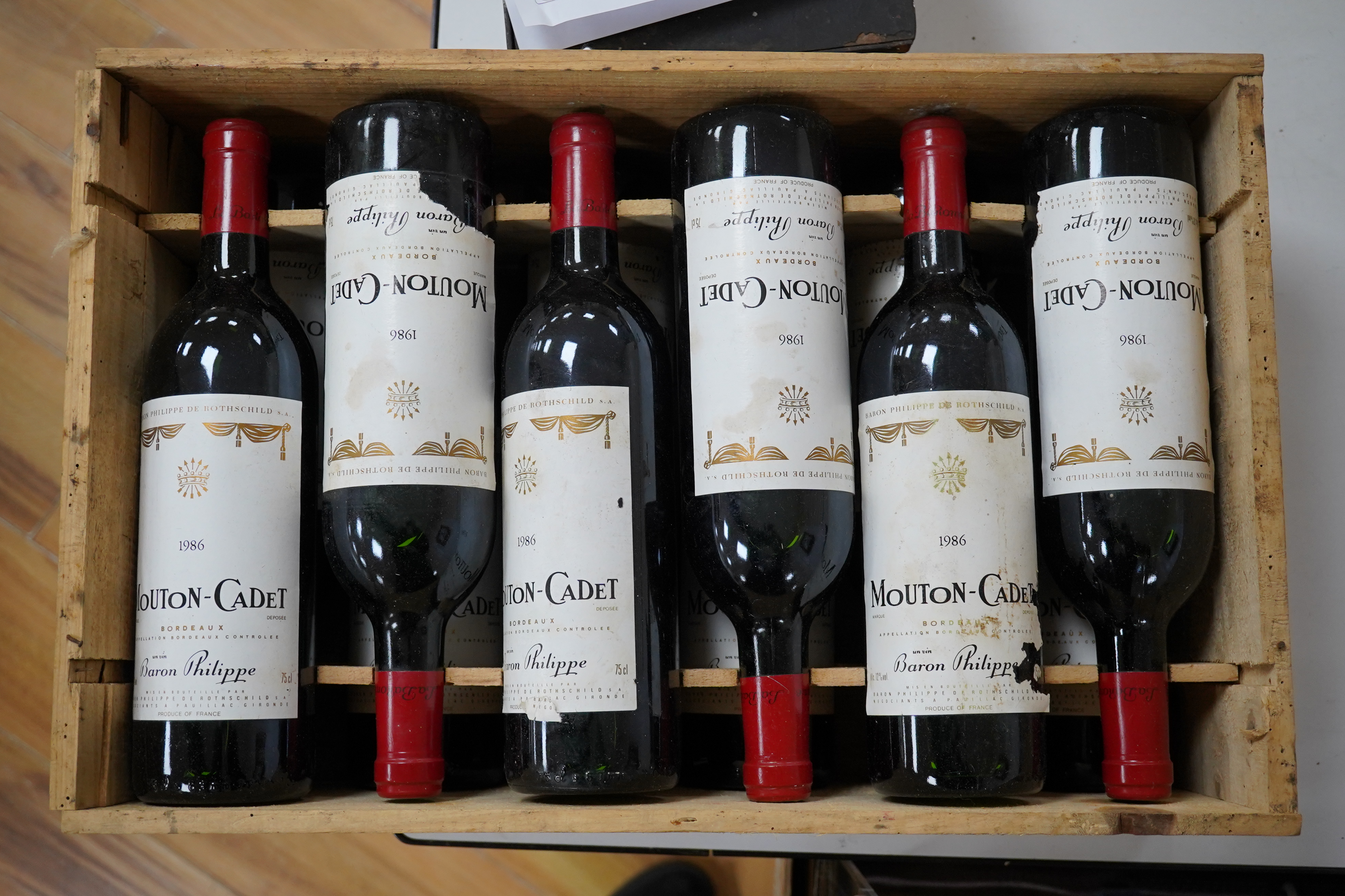 Twelve bottles of Mouton Cadet 1986 in associated wooden crate. Condition - wear to labels, storage unknown